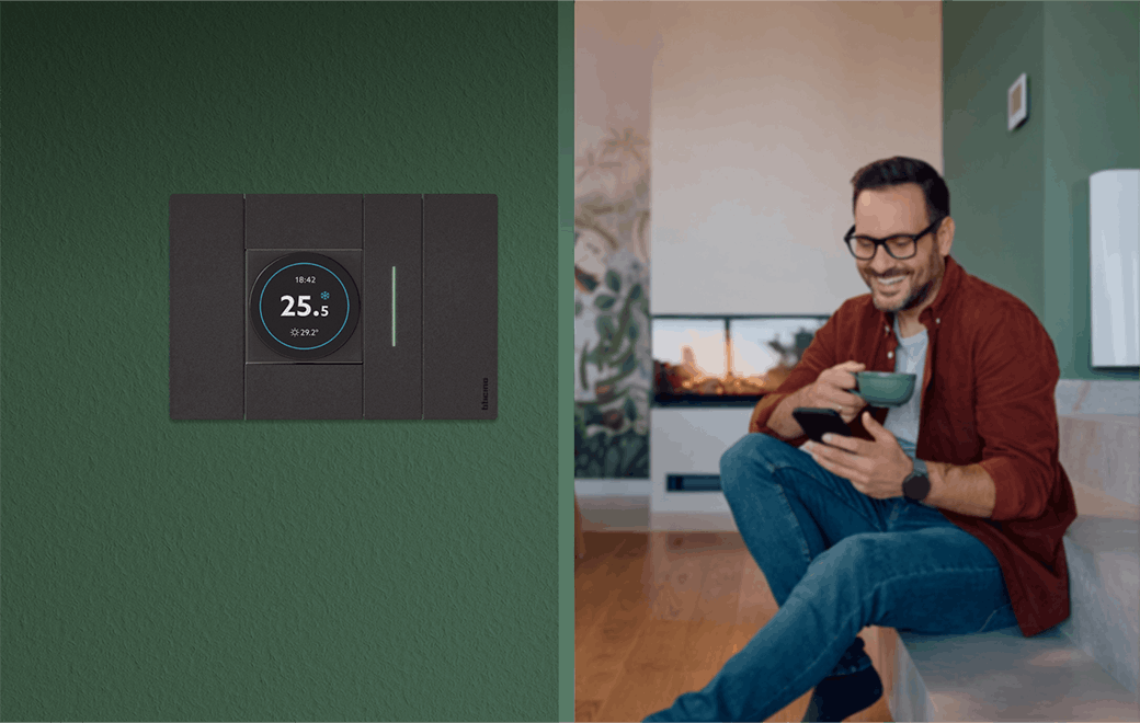 Swipe di 4box installato su una parete verde, con display OLED attivo, mentre l’utente gestisce da smartphone le funzioni di controllo remoto del comfort domestico.